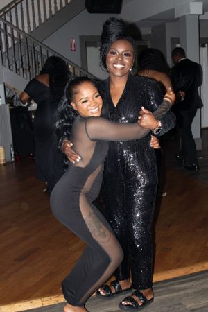 Two friends in formal black attire sharing a joyful moment during a party.