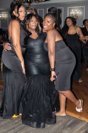 Three women in elegant black dresses pose together at a formal event on hardwood floors.