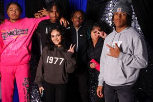 Group posing together wearing casual streetwear including hoodies and sweatshirts against dark background with blue lighting.