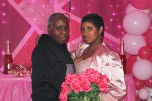 Couple posing with pink roses at a Valentine's Day party with pink backdrop and white balloons.