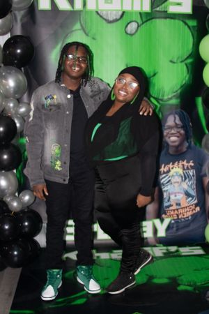 Two people posing in front of green backdrop with black and silver balloons at an event.