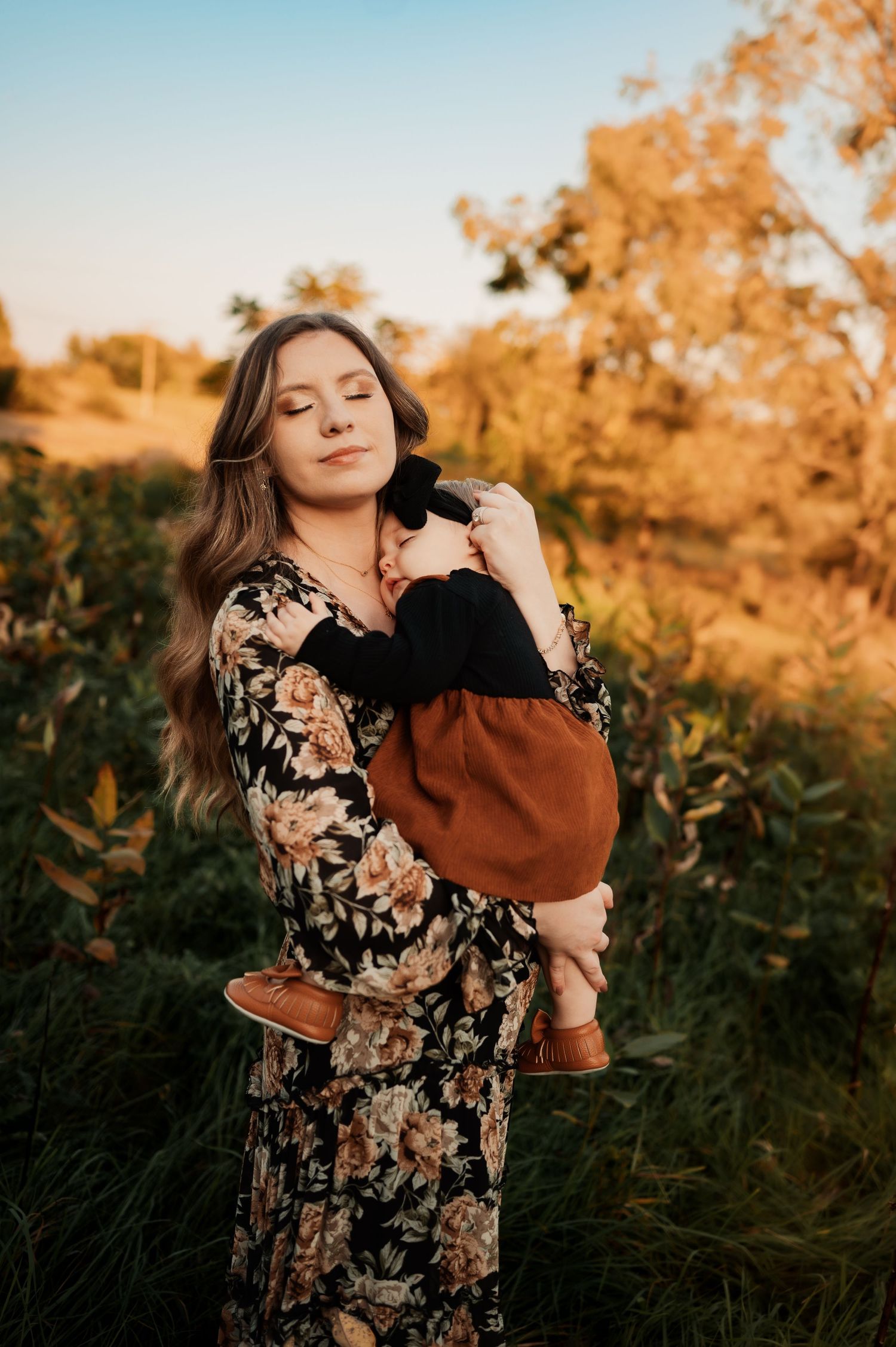 Woman in floral dress holds small black cat while standing in golden sunlit field.