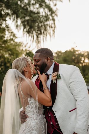 Bride and groom sharing a tender kiss outdoors, groom in white tuxedo with burgundy vest and boutonniere.