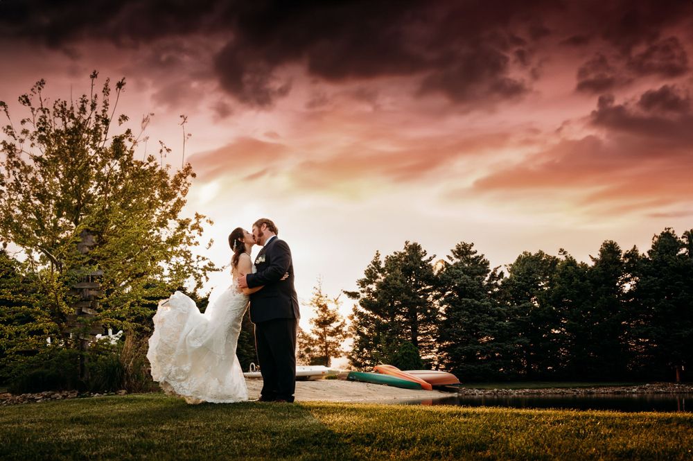 Groom dips bride in white gown under a dramatic sunset sky in a stunning wedding photo.