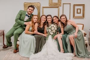 Bride in white gown poses with bridesmaids and groomsman in sage green attire indoors.