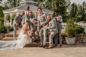 Groomsmen in gray suits celebrate with the groom and bride near a white gazebo at an outdoor wedding.