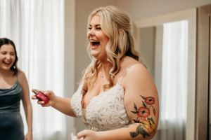 Laughing bride with floral arm tattoo reacts joyfully while getting ready on wedding day.