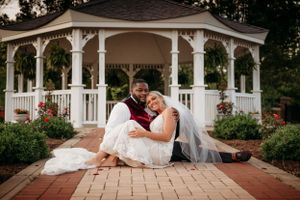 Bride and groom sitting together in front of a white gazebo surrounded by roses in a garden.