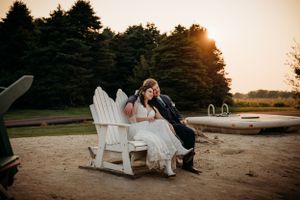 Bride and groom sit together on white Adirondack chairs during a golden sunset at their wedding.