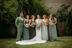 Bride and wedding party in sage green dresses and suit smile outdoors in a garden setting.