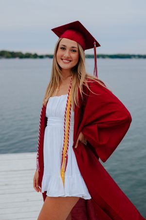 Graduate in red cap and gown poses on waterfront dock during sunset celebration.