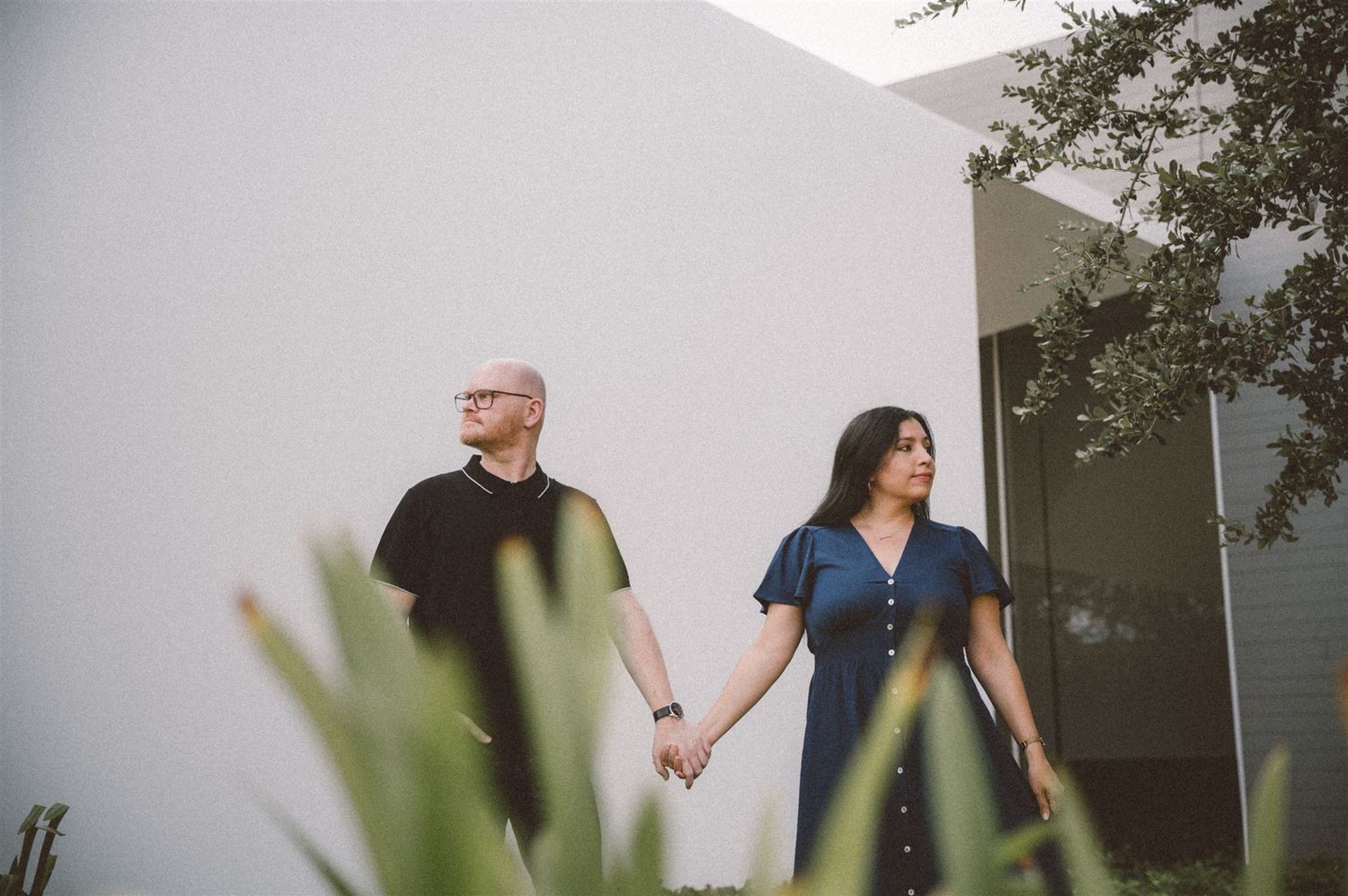 A couple walks hand in hand past a modern white building with plants in the foreground.