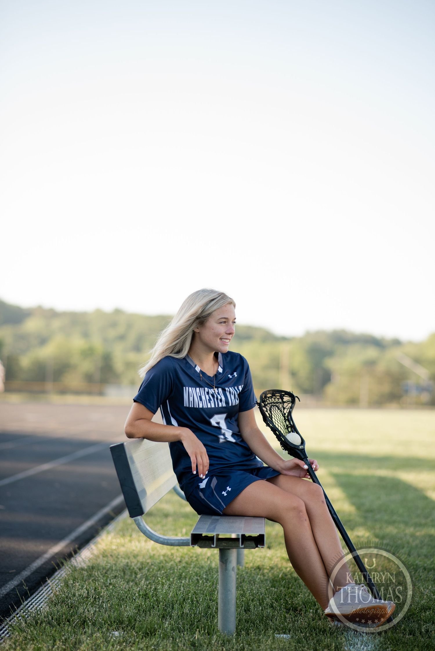 Student athlete rests on bench with lacrosse stick at outdoor sports field.