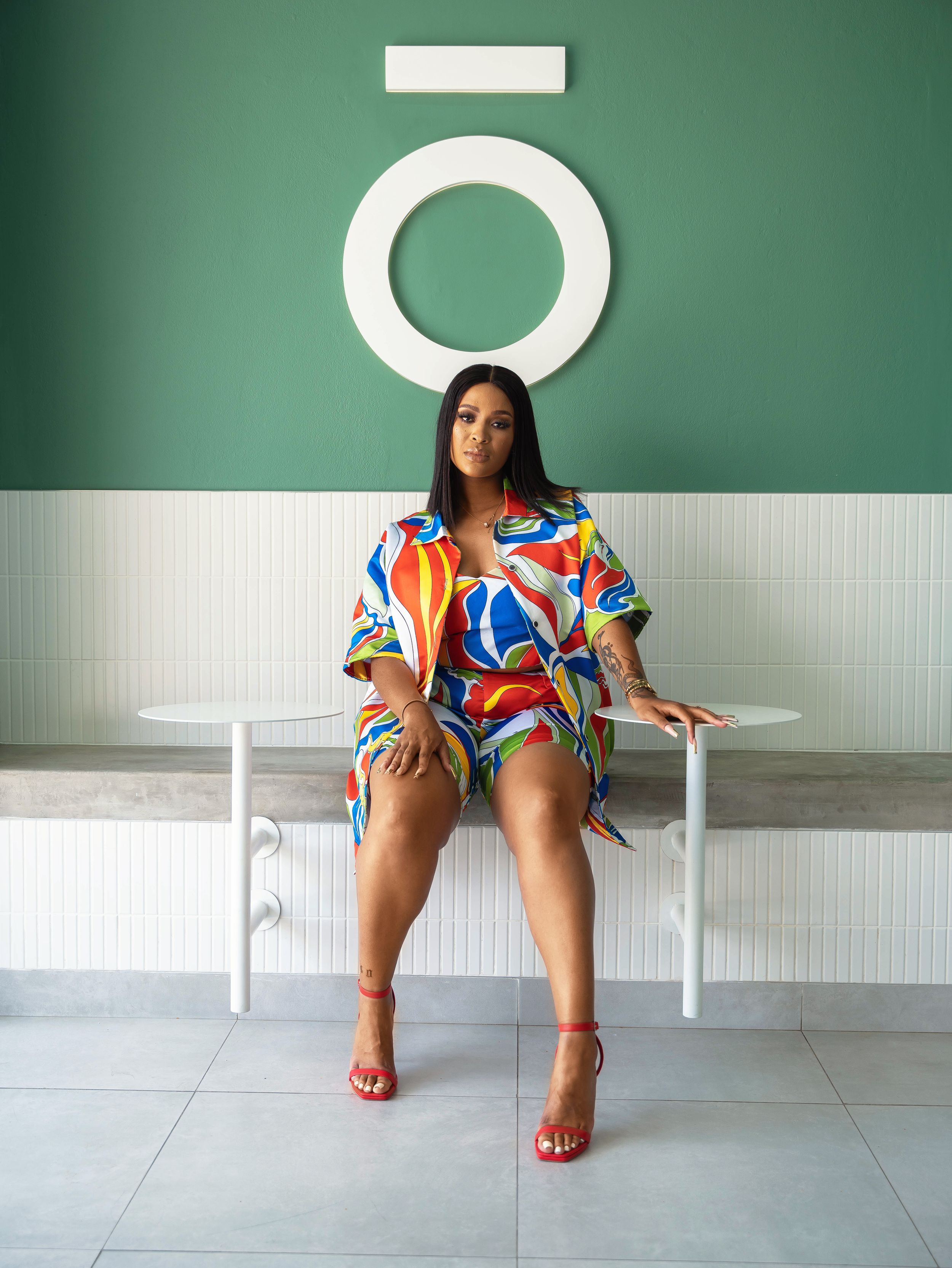 Model in colorful printed outfit sits on white bench against green wall with circular decor.