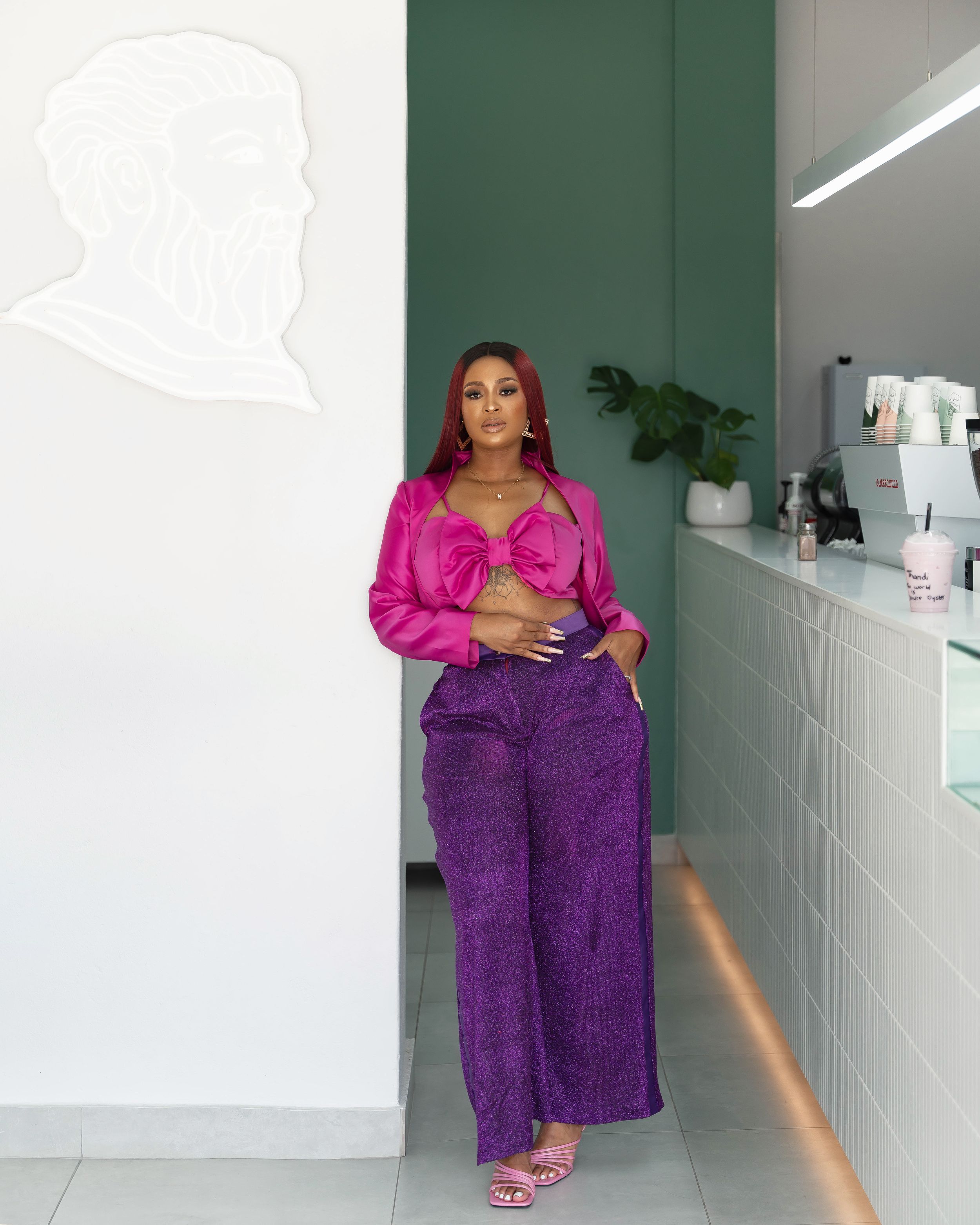 Fashion model wearing fuchsia top and purple pants stands elegantly in minimalist hallway setting.
