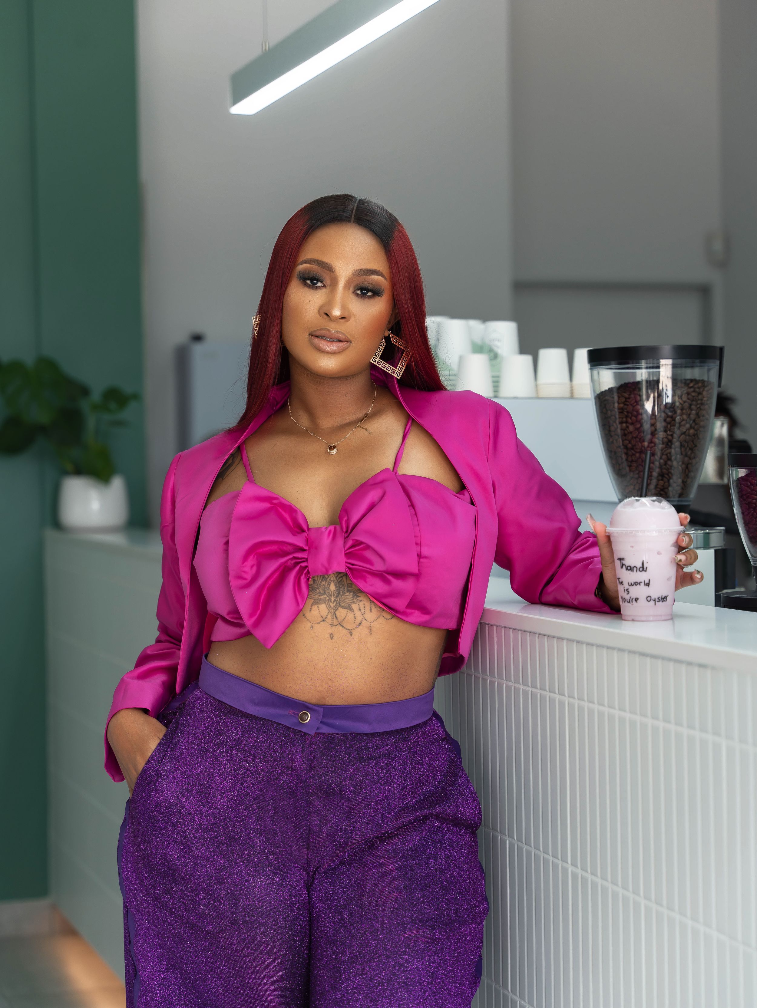 Model in bright pink crop blazer and purple pants poses confidently in modern interior setting.