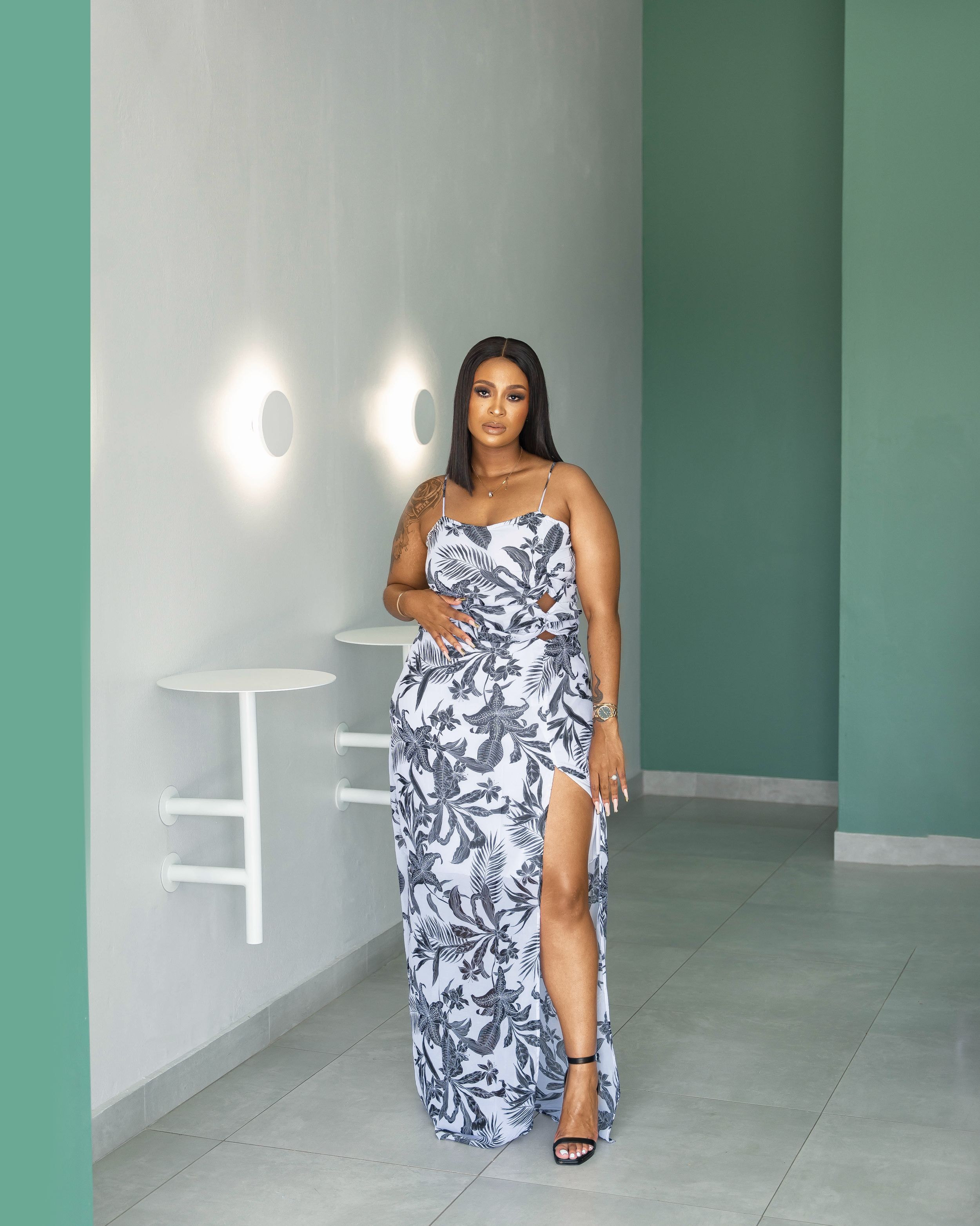 Model in floral print maxi dress with thigh slit poses elegantly against mint green wall with sconce lighting.