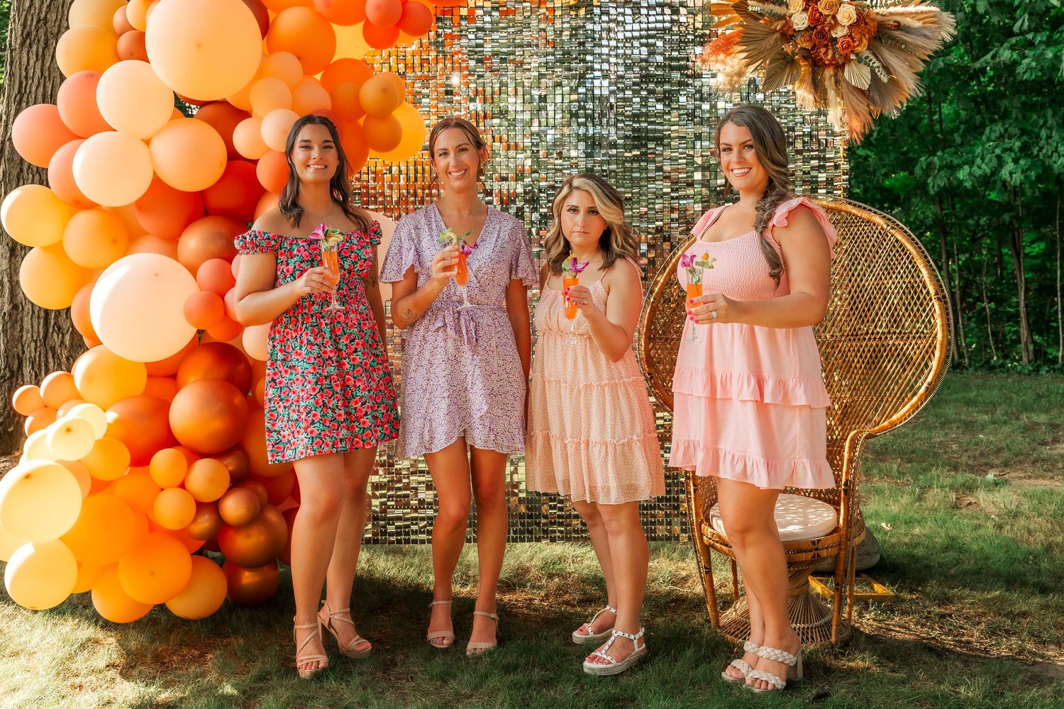 Group celebrating at outdoor party with orange balloon arch and peacock chair.