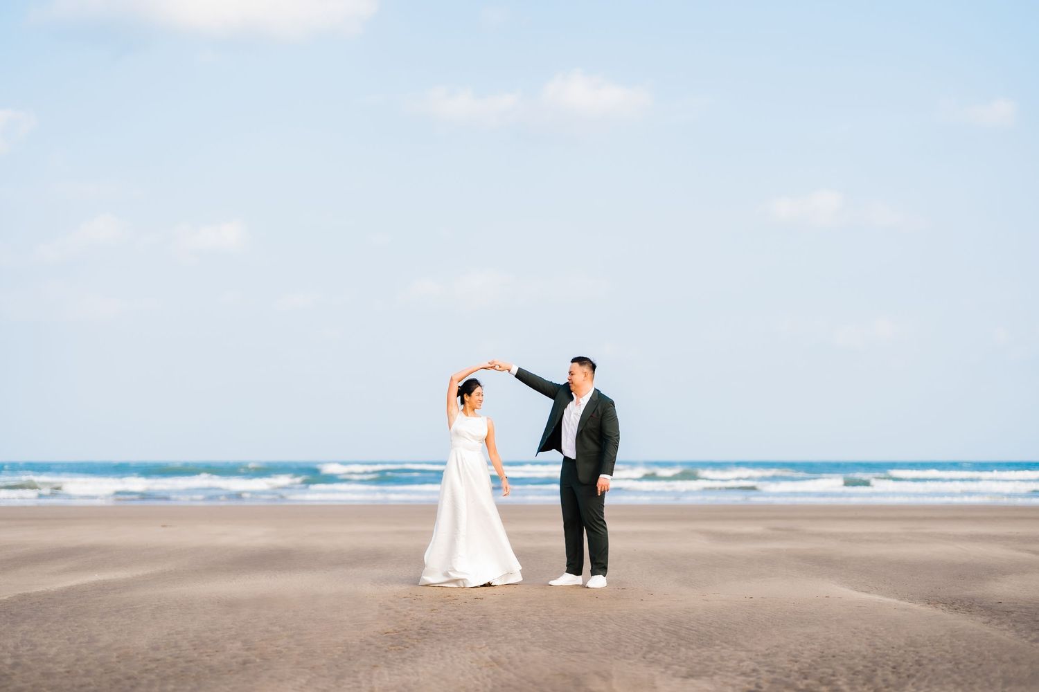 A couple in wedding attire standing together on a sandy beach with ocean waves in the background.