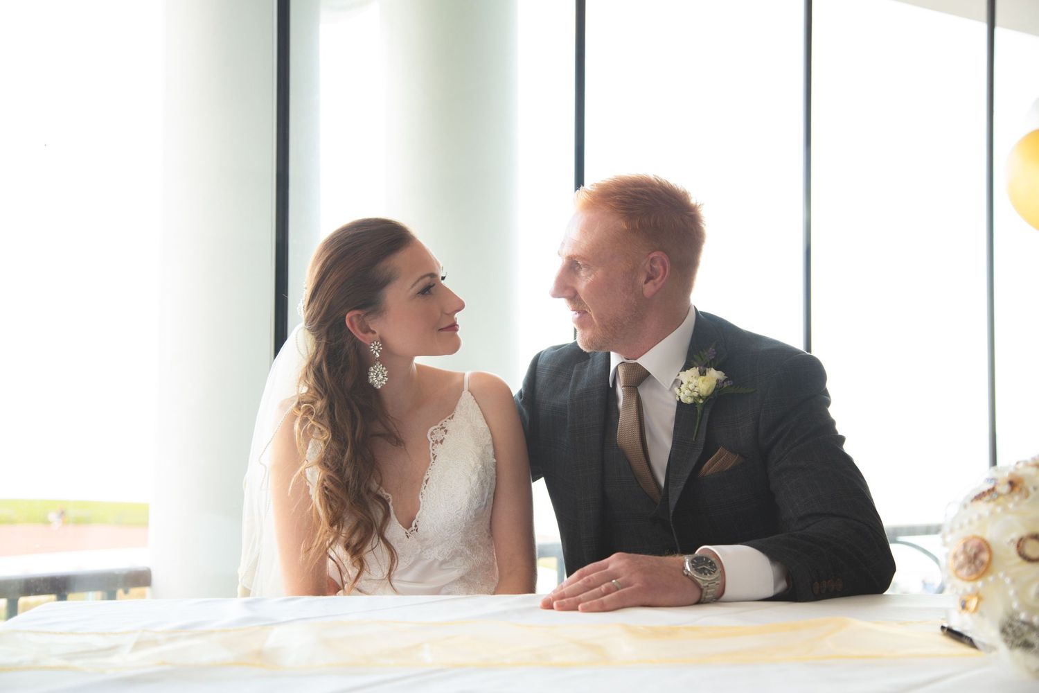 Wedding couple sharing intimate moment at reception table by bright windows.
