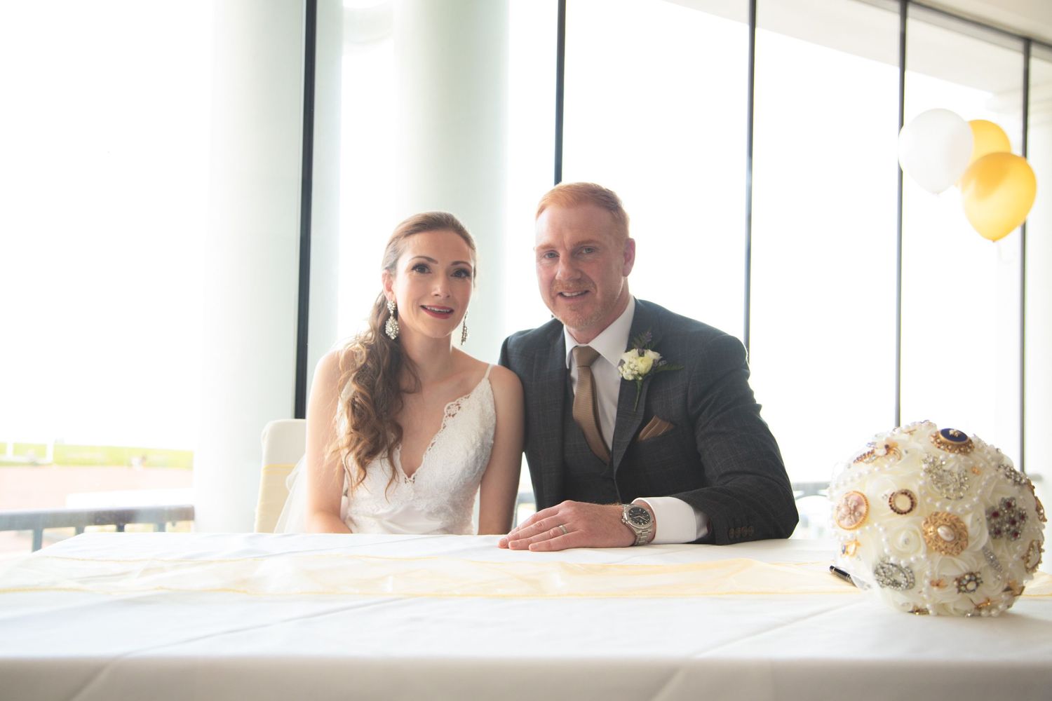 Wedding couple sitting at table with decorative bouquet in bright modern venue.