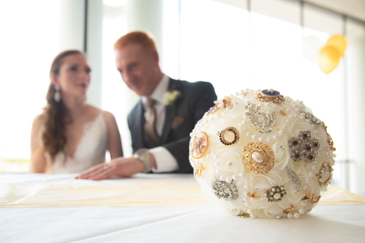 Ornate white and gold brooch bouquet in focus with wedding couple blurred in background.
