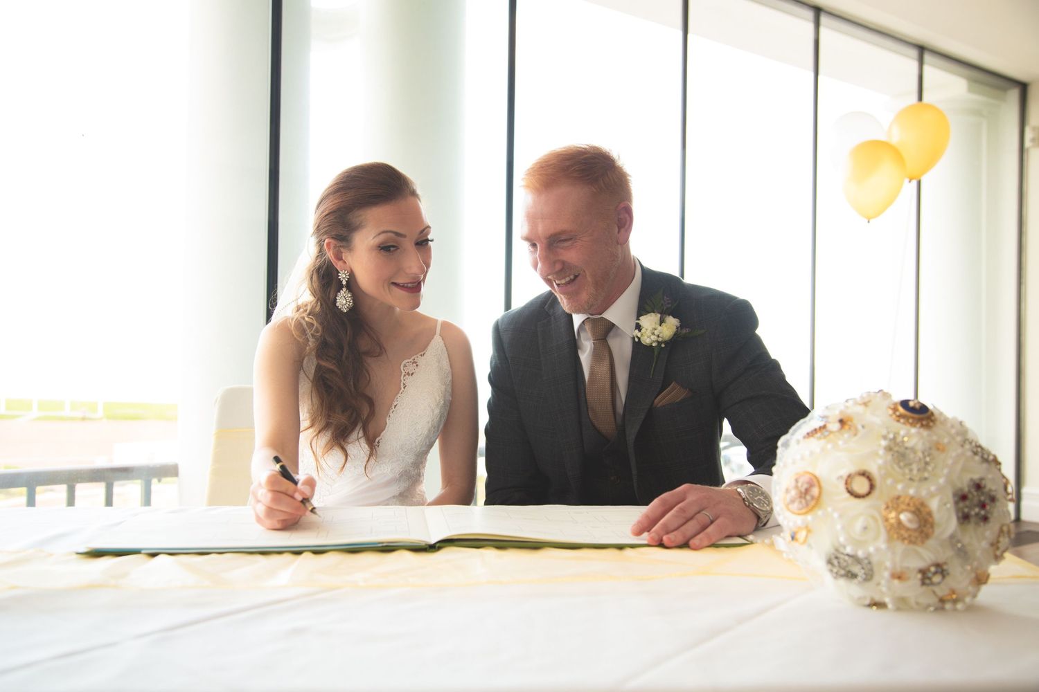 Couple signing documents at bright wedding venue with decorative brooch bouquet on table.