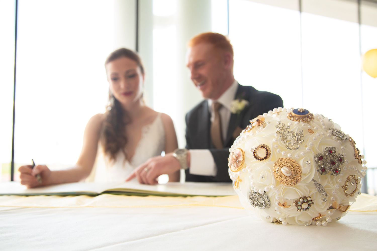 Wedding couple signs marriage documents with elegant pearl and crystal brooch bouquet in foreground.
