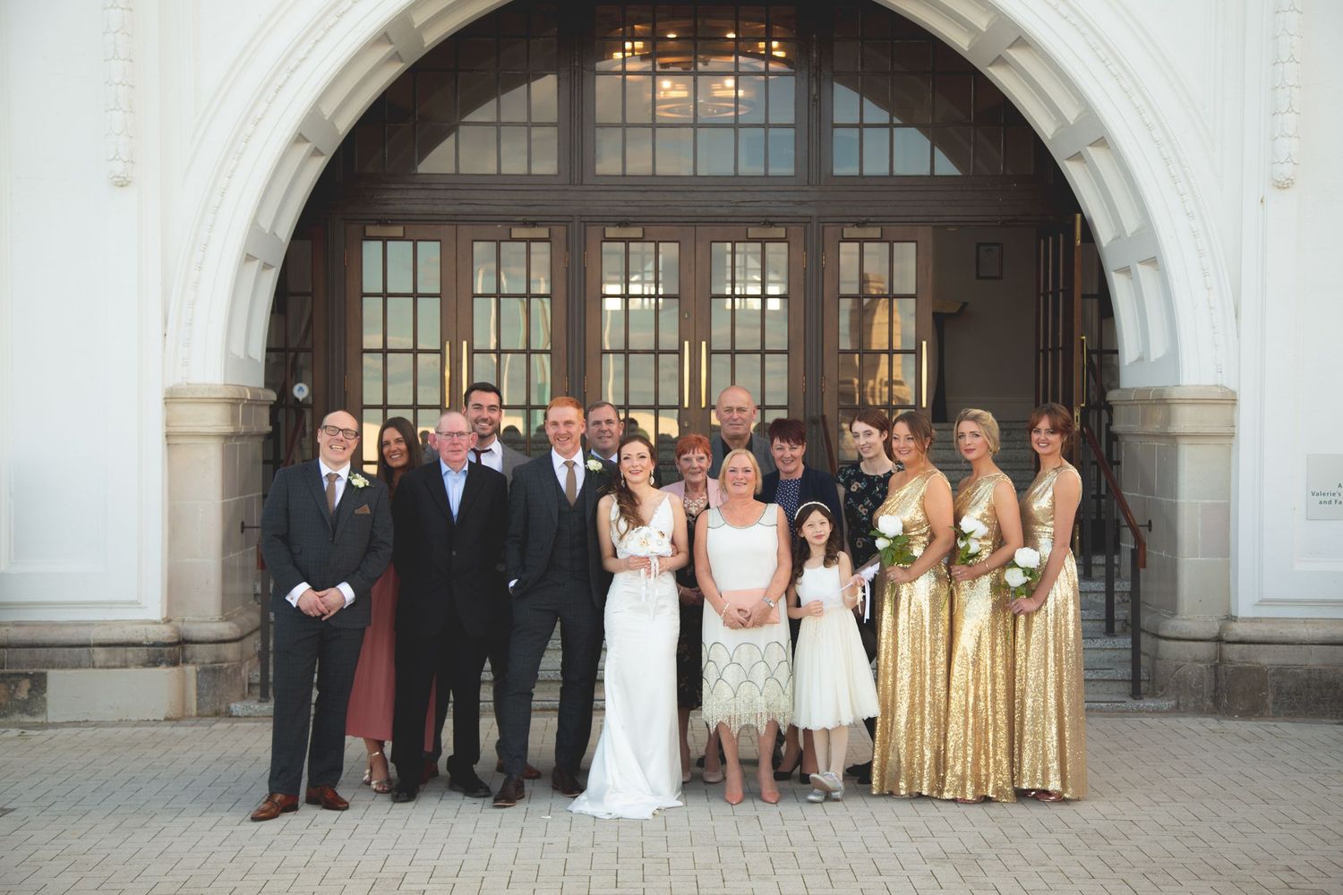 Wedding party poses together under large arched entrance of historic building.