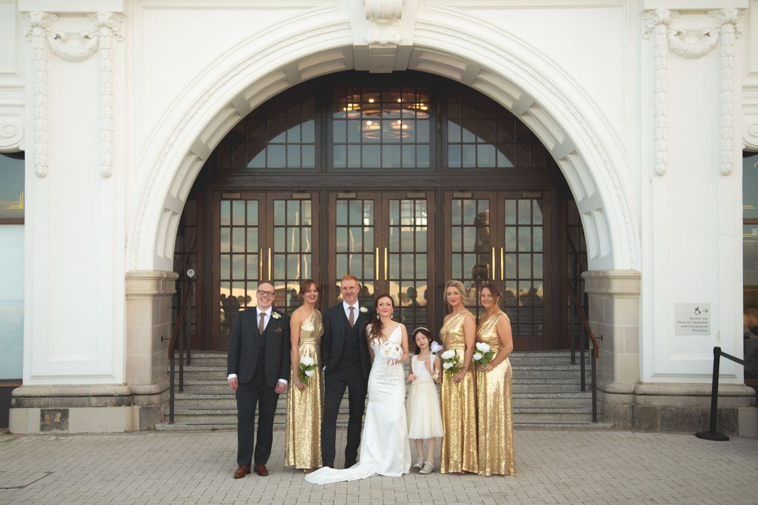 Wedding party in gold and formal attire posed before classic arched entrance.