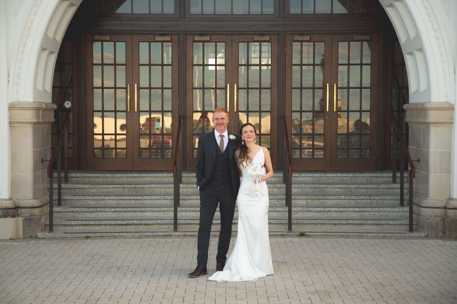 Romantic wedding portrait of couple in formal attire standing before grand arched entrance.