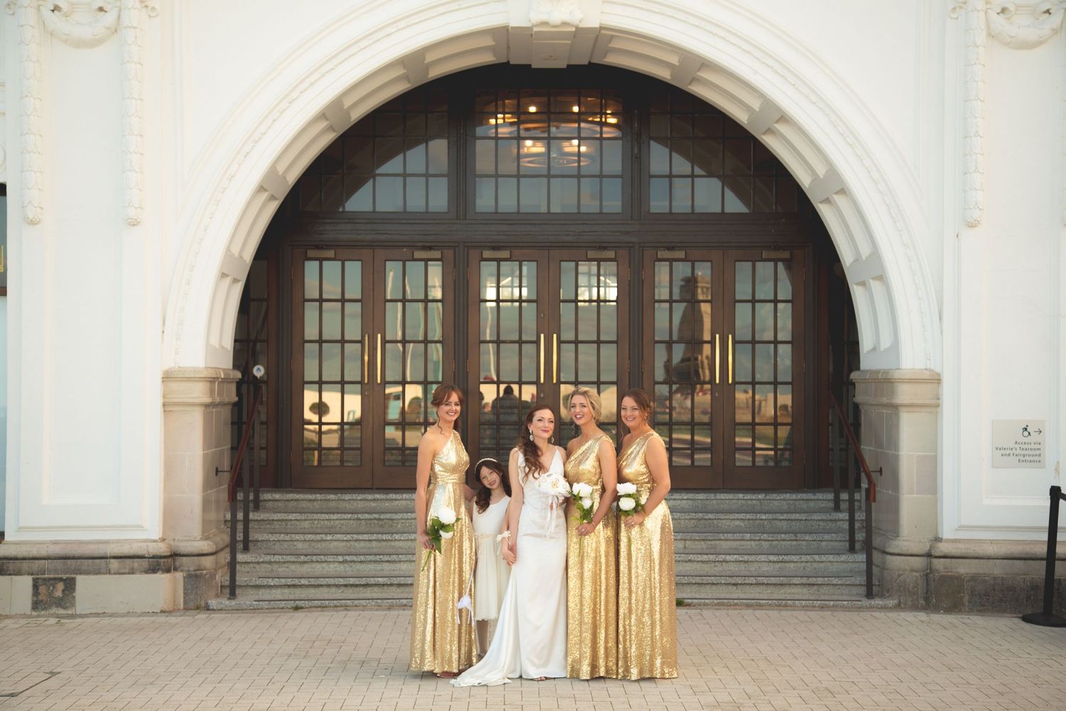 Wedding party in gold colored dresses posed in front of elegant arched doorway.