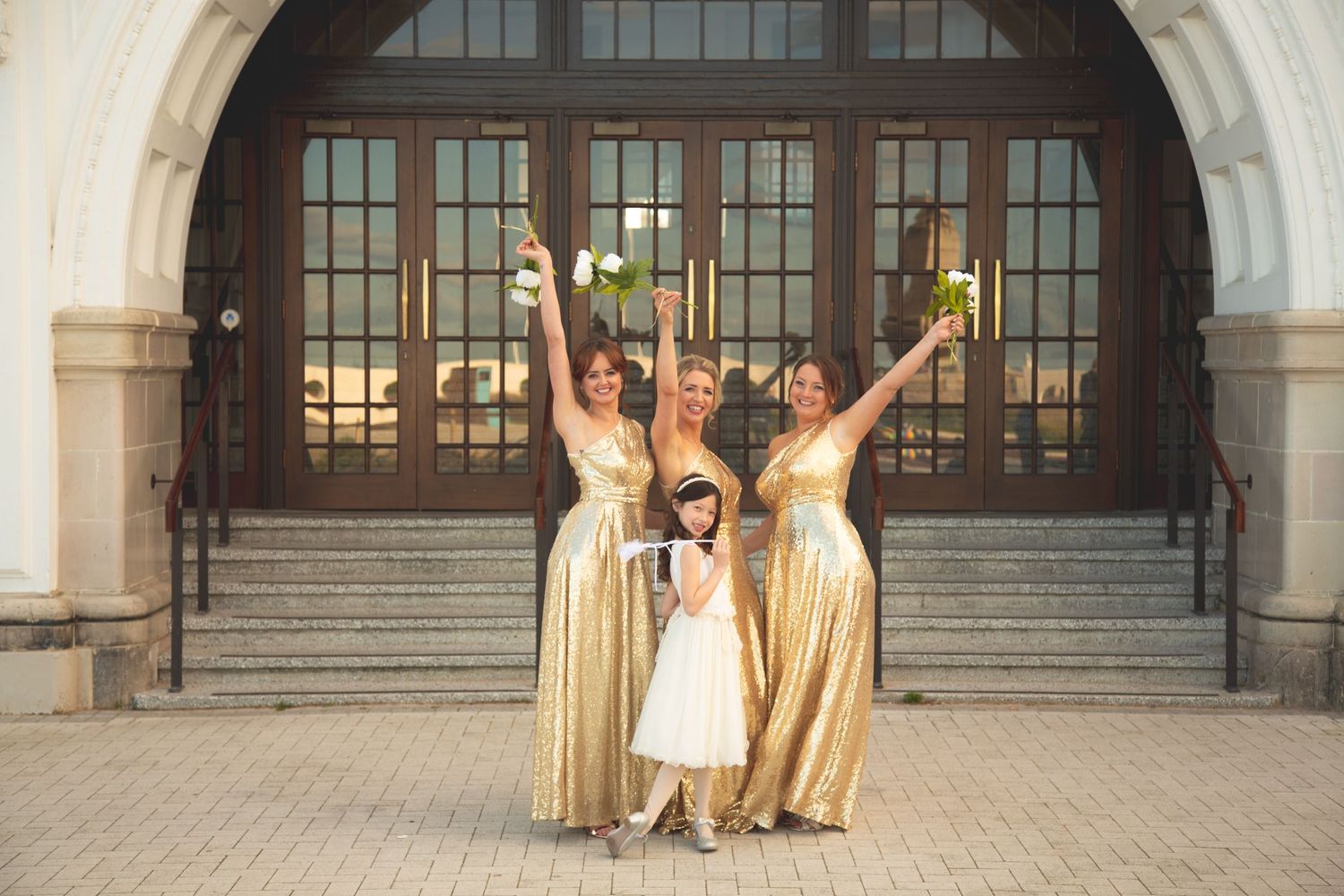 Wedding party in gold dresses celebrating joyfully on steps of building with arched entrance.