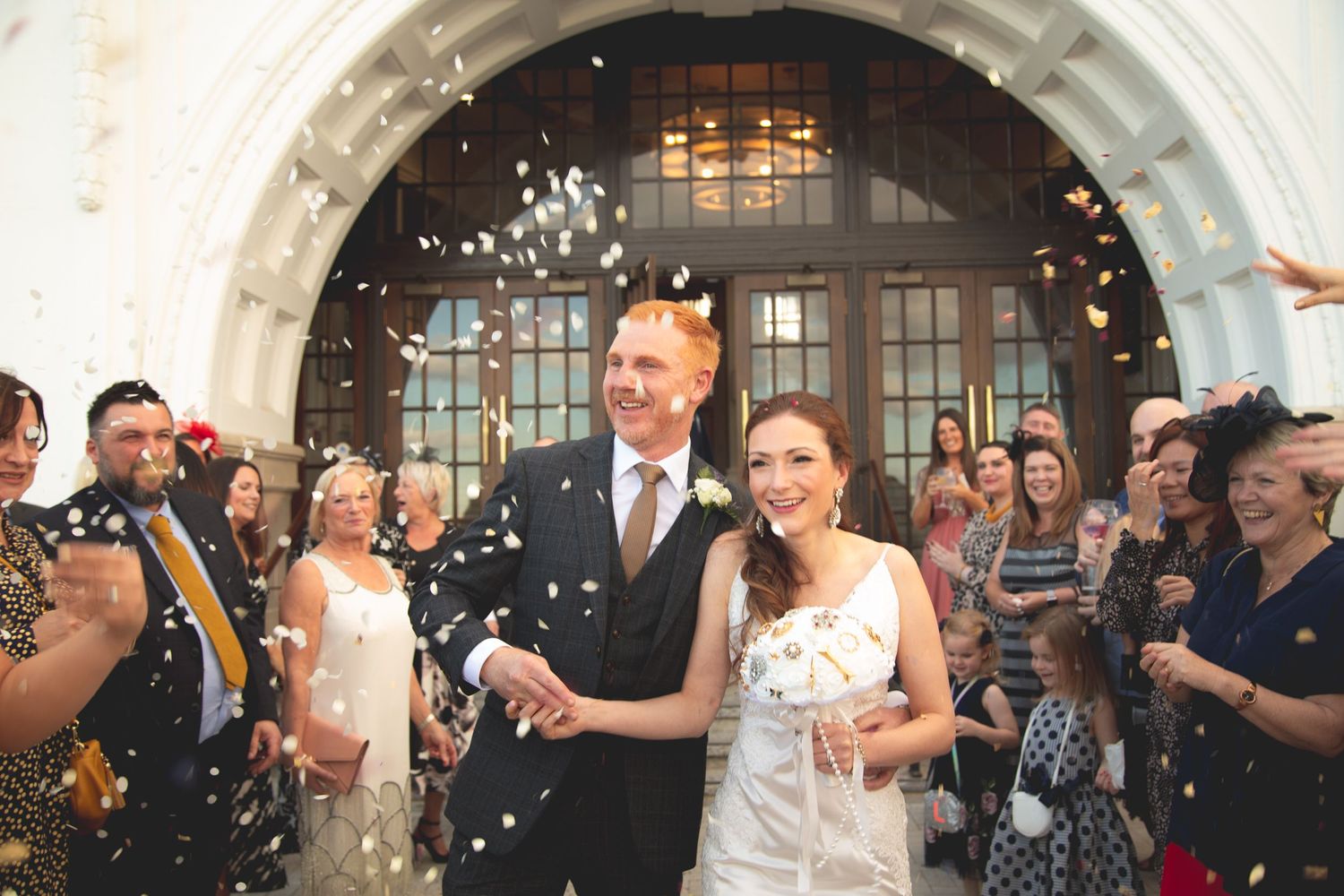 Newlyweds walk through shower of confetti with guests cheering outside arched doorway.