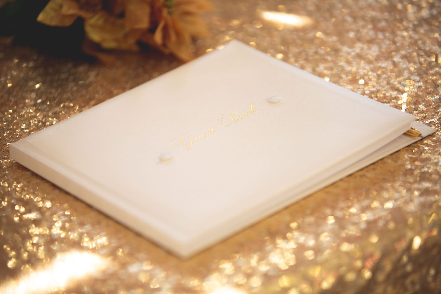 A white guest book sits on a shimmering gold sequined surface.