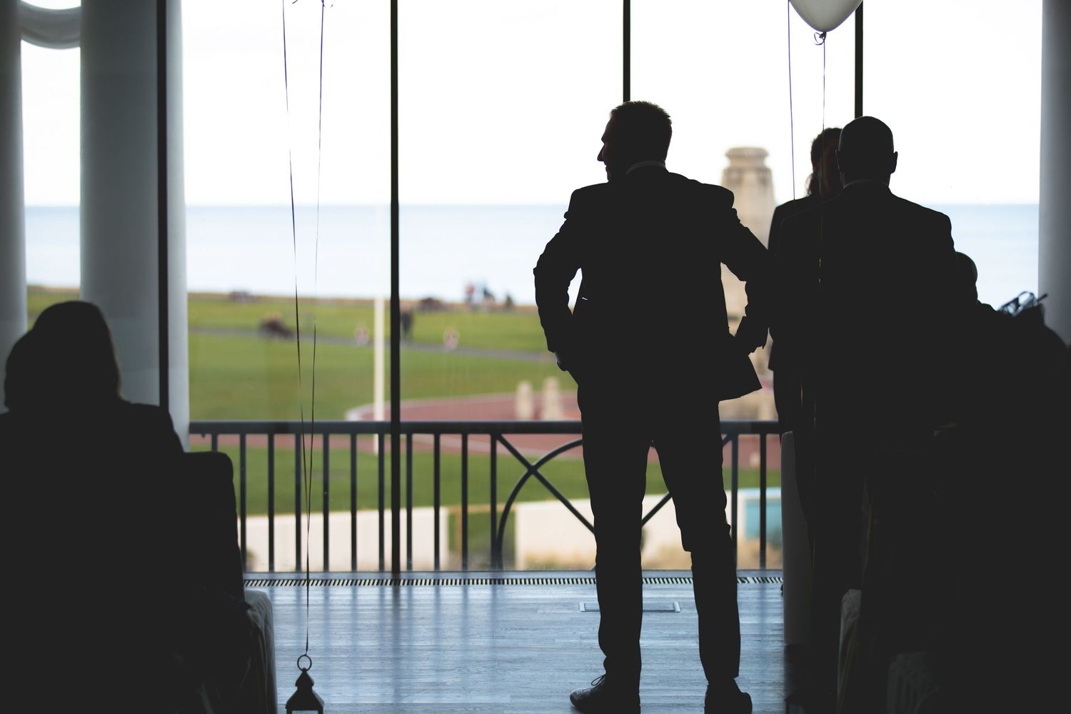 Silhouettes of wedding guests stand against large windows overlooking a scenic landscape.