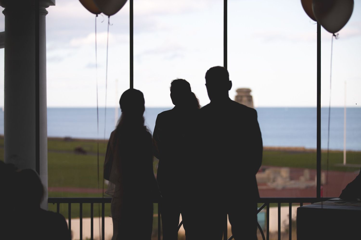 Silhouettes of wedding guests gather by large windows with balloons.