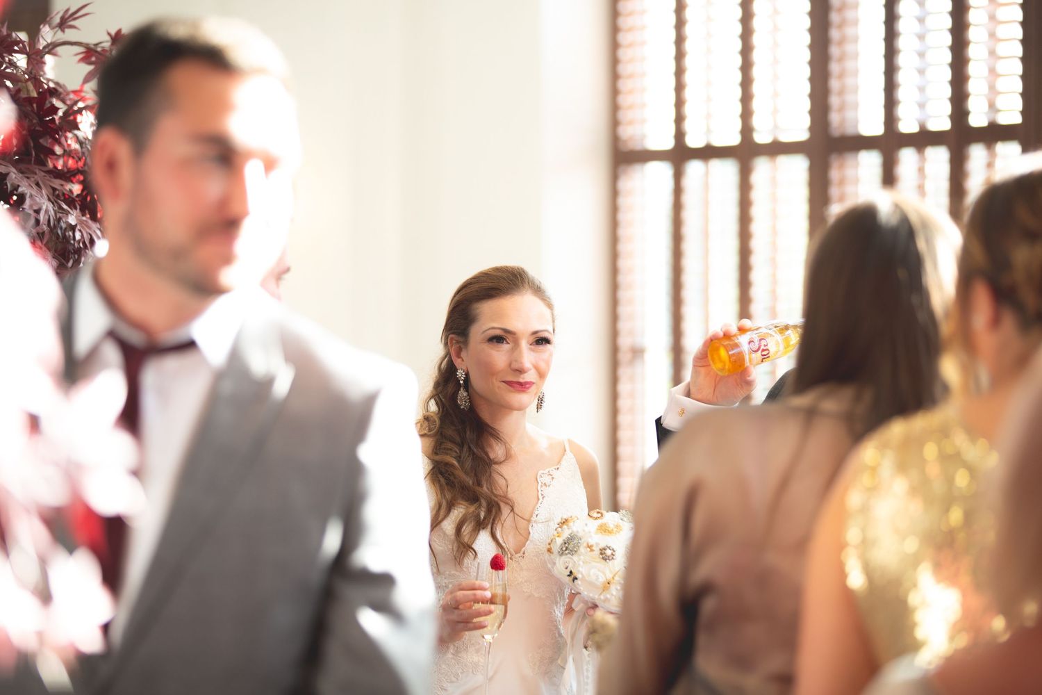 Wedding guests mingle and socialize during an elegant reception with sunlit windows in the background.
