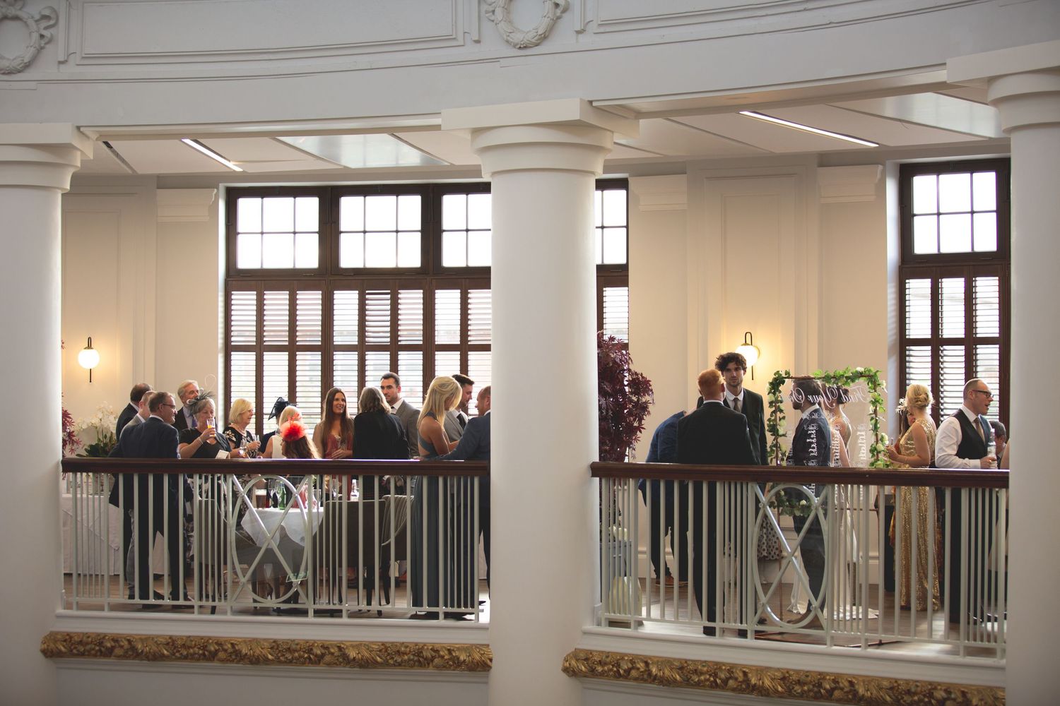 Wedding guests gathered on an elegant mezzanine level with large windows and ornate architectural details.