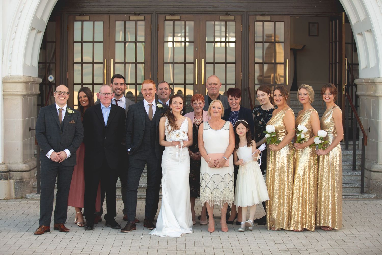 Wedding party poses for formal group photo in front of grand arched entrance with ornate windows.