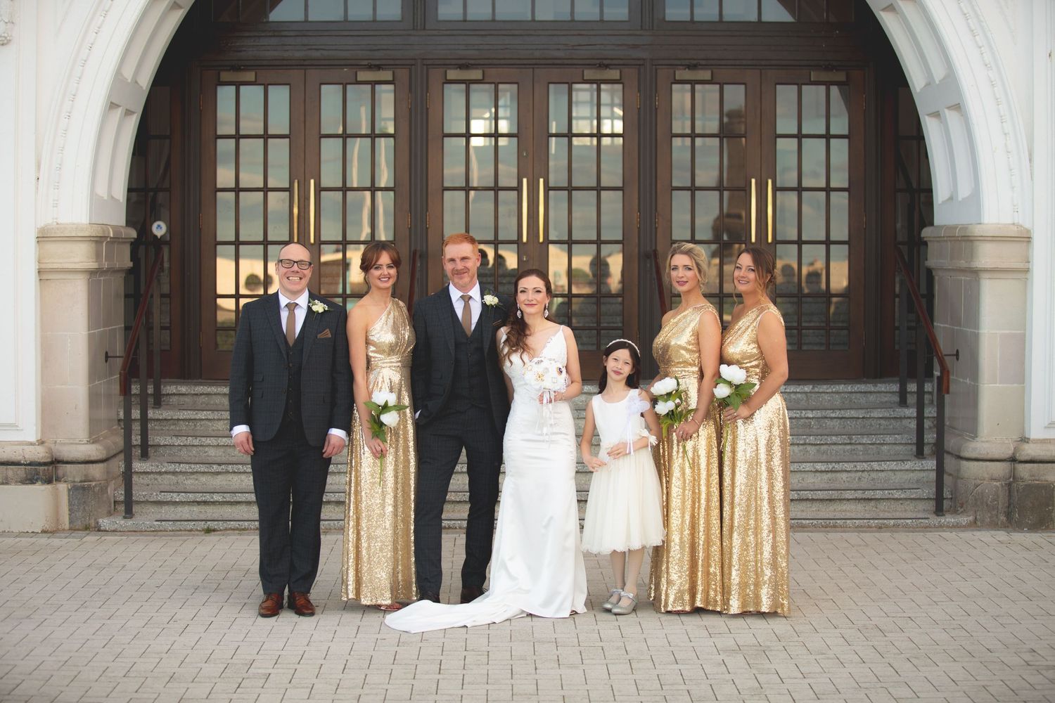 Wedding party in gold dresses and formal attire posed on stone steps under arched entrance.