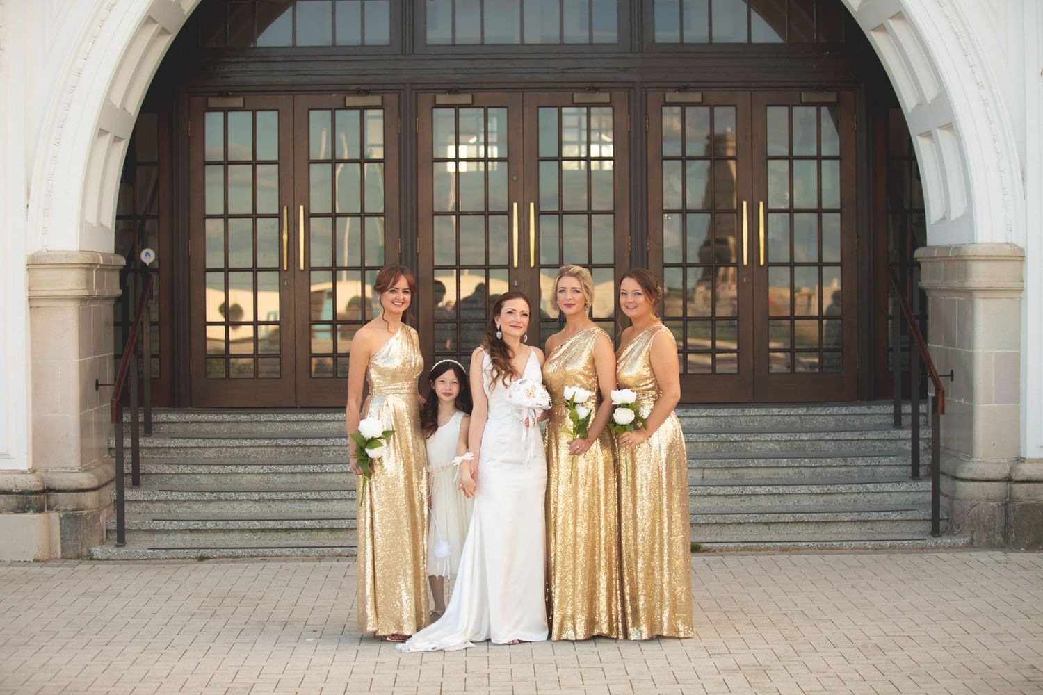 Bride in white gown poses with bridesmaids in gold dresses on stone steps.
