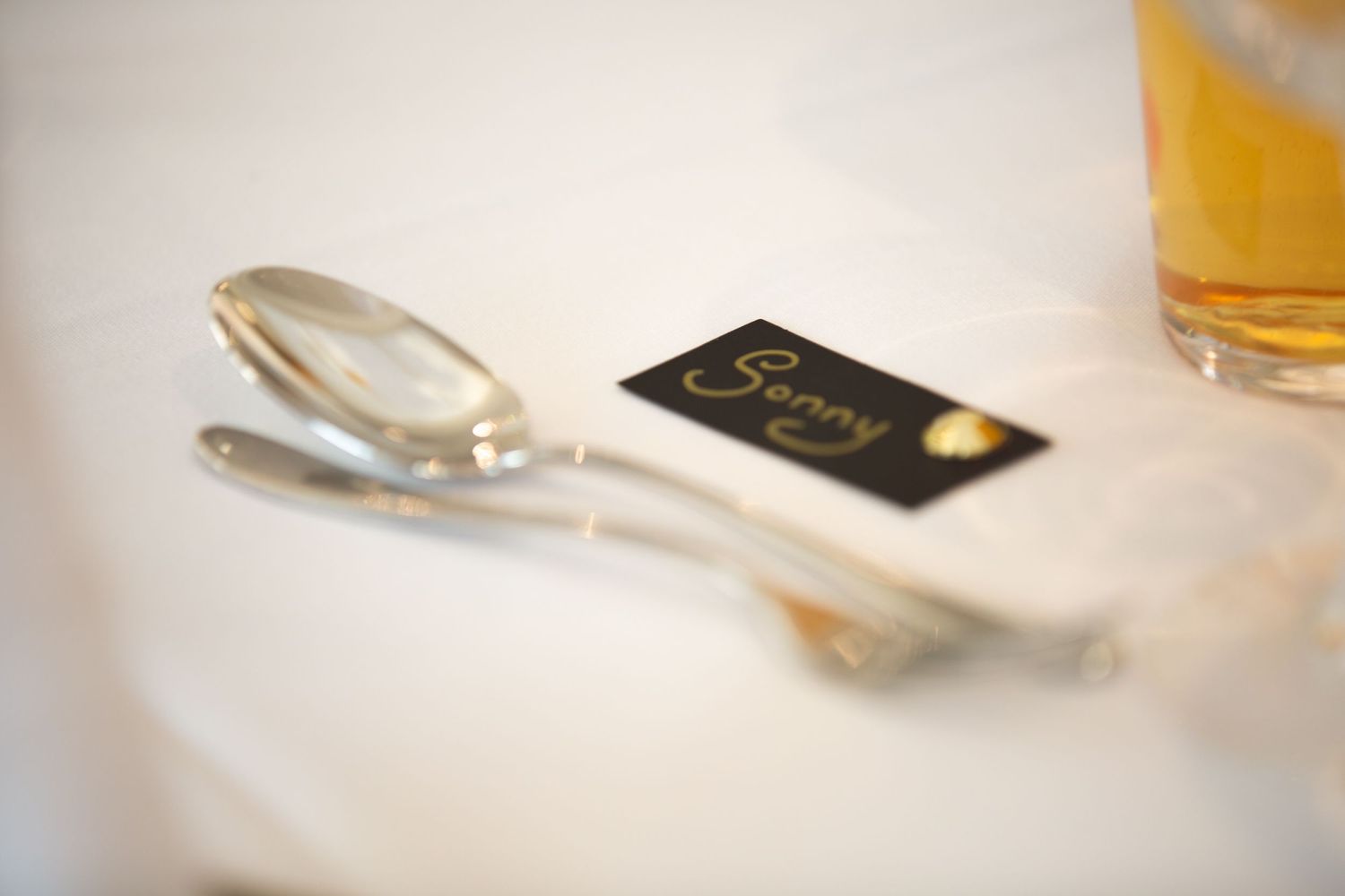 Place setting detail with silver cutlery and black name card on white tablecloth.