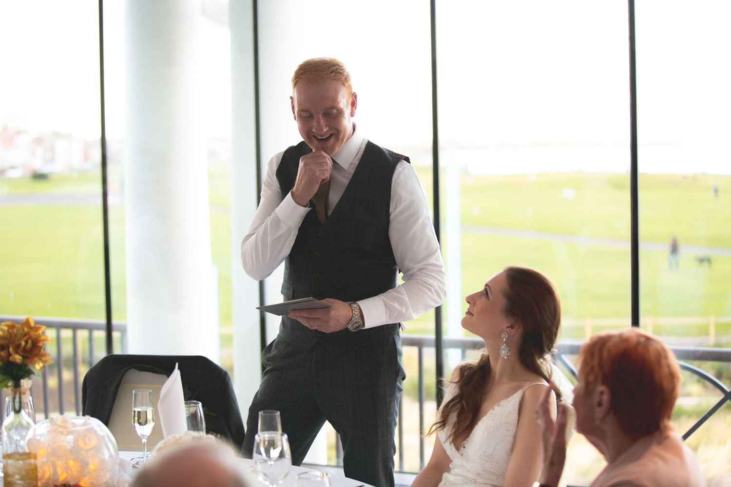 A speaker in formal attire addresses guests during a wedding reception by large windows.