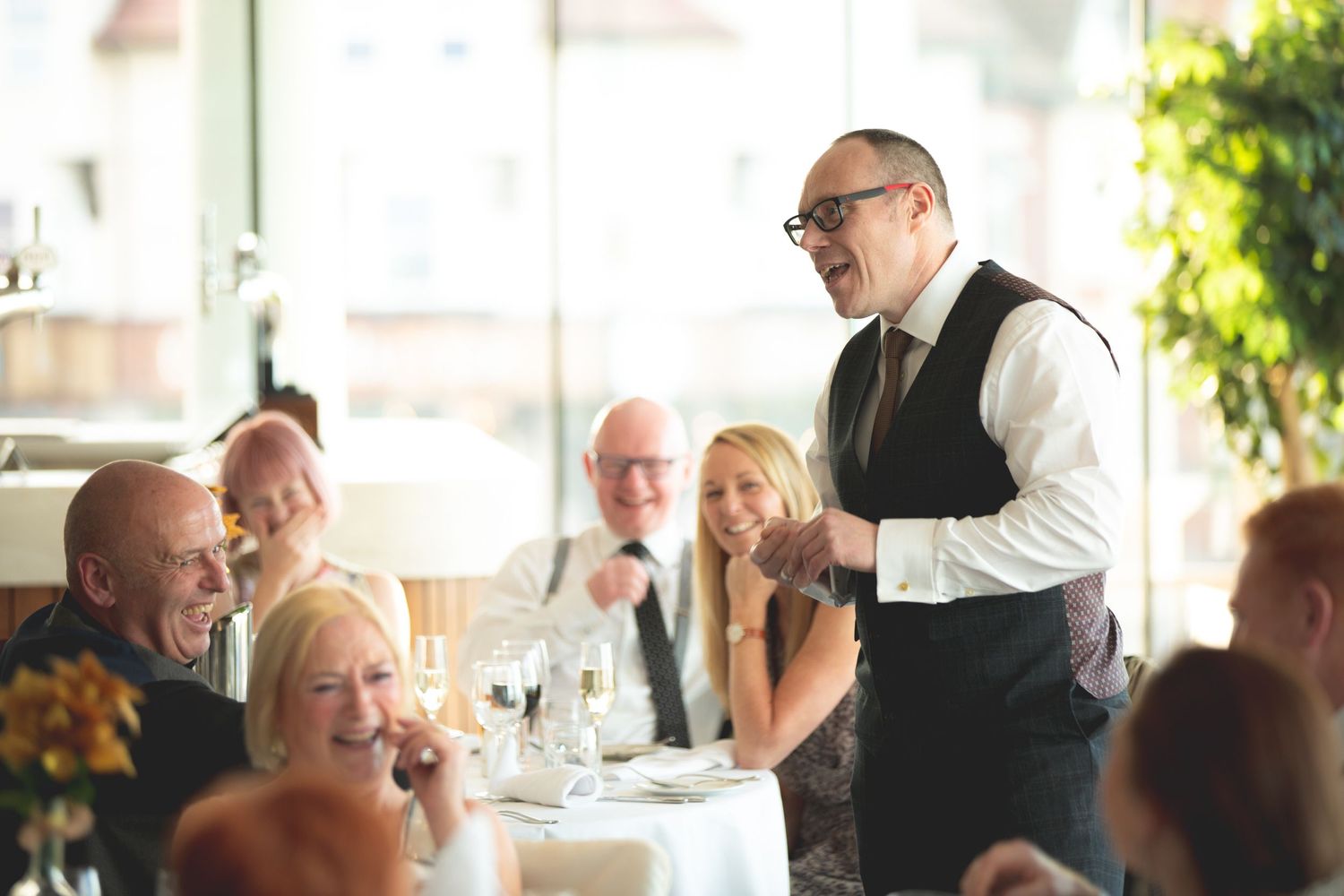 Elegant dinner party scene with server and guests interacting at white-clothed tables.