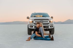 Couple sitting together in front of pickup truck on salt flats at sunset.