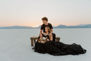 Romantic couple in formal attire poses on bench at salt flats during sunset with mountains in background.
