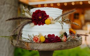 White wedding cake decorated with burgundy dahlias and yellow flowers on rustic wood stand.