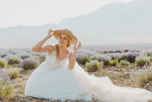 A bride in a flowing dress and sun hat sits among desert lavender fields at sunset.