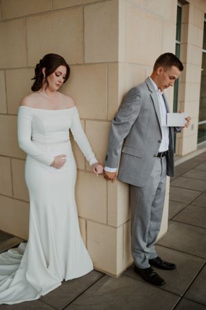 A couple in wedding attire holds hands around a corner in a first look moment.